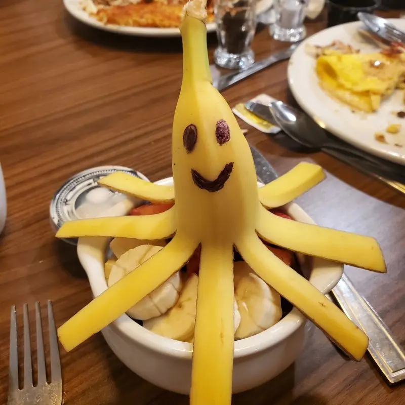 A banana cut into the shape of an octopus with a smiley face drawn on sits atop a bowl of fruit.
